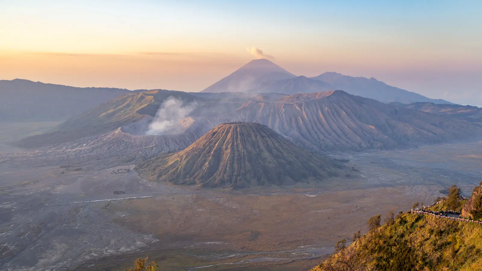 The volcanic cones of Mount Bromo and the Tengger caldera under a soft dawn sky, with a plume rising from distant Semeru